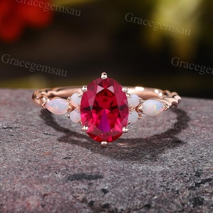 May include: Rose gold ring featuring a large oval-cut ruby center stone, flanked by small white opal accents. The ring's band has a delicate, textured design. The ruby is a vibrant red, and the opals have a milky white appearance. The ring is set on a dark, textured surface.