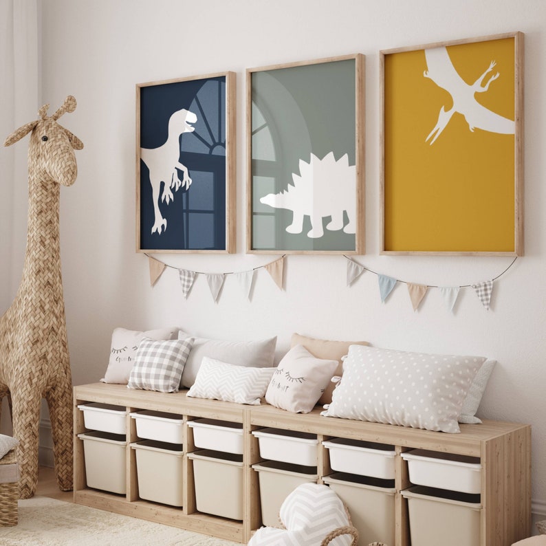 May include: Three framed prints with white dinosaur silhouettes on colored backgrounds. The prints are hung on a white wall above a wooden bench with storage bins. The bench has a light wood finish and is decorated with pillows.