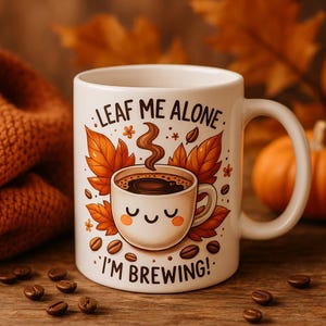 May include: White ceramic coffee mug with a whimsical design. The mug features a smiling coffee cup illustration surrounded by autumn leaves and coffee beans. Text on the mug reads "LEAF ME ALONE I'M BREWING!" The mug has a curved handle and is set against a blurred background.