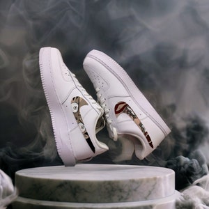 Puede incluir: Un par de zapatillas de cuero blanco con ilustraciones personalizadas en el logotipo de Nike. La ilustración presenta un personaje de dibujos animados con ojos grandes y una combinación de colores rojo y marrón. Las zapatillas se exhiben sobre un pedestal de mármol sobre un fondo ahumado.