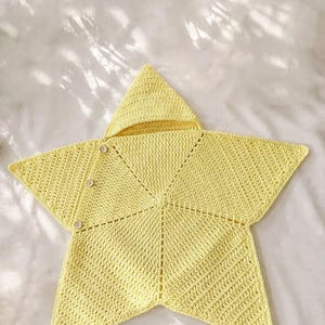 May include: A pale yellow star-shaped crochet blanket featuring a pointed hood at the top. The garment is constructed with a textured stitch pattern and includes three small, round, light-colored buttons along one of the side points for closure.