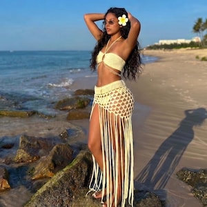 May include: A woman's beach outfit, featuring a crochet top and skirt with fringe. The outfit is a light yellow color. The background includes a beach, ocean, and blue sky.