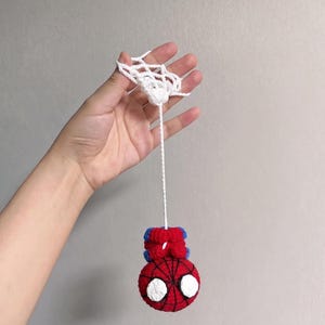 May include: A crocheted red and blue spherical ornament featuring a black web pattern and large white eyes, suspended by a thin white cord from a white crocheted web-like structure. The item is crafted with yarn in a round, plush design.