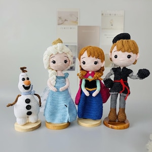 K&ouml;nnte beinhalten: Geh&auml;kelte Puppen von Charakteren aus dem Animationsfilm Frozen. Die Puppen umfassen Olaf den Schneemann, Elsa in einem blauen Kleid, Anna in einem blau-rosa Kleid und Kristoff in einem schwarzen Outfit. Jede Puppe steht auf einem Holzsockel.