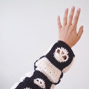 May include: A long-sleeved crochet garment featuring a repeating pattern of black and white squares. Each square contains a white peace sign motif centered within a black border, creating a high-contrast graphic design across the fabric.