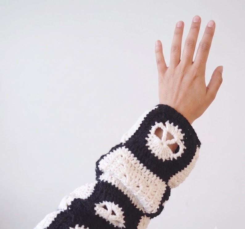 May include: A long-sleeved crochet garment featuring a repeating pattern of black and white squares. Each square contains a white peace sign motif centered within a black border, creating a high-contrast graphic design across the fabric.