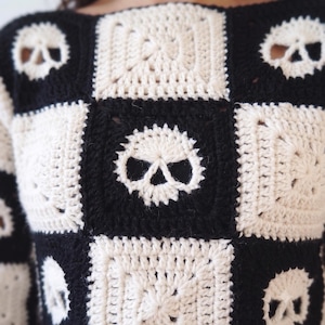 May include: A long-sleeve sweater constructed from alternating black and off-white crocheted granny squares. The design features a repeating pattern of skull motifs integrated into the squares, creating a checkerboard effect across the garment.