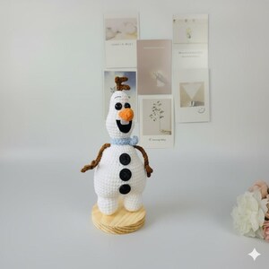 K&ouml;nnte beinhalten: Eine geh&auml;kelte Olaf-Schneemannfigur mit Karottennase, schwarzen Knopfaugen und einem blauen Schal. Der Schneemann hat braune Zweigarme und einen Holzsockel. Die Figur steht vor einem wei&szlig;en Hintergrund mit dekorativen Karten.