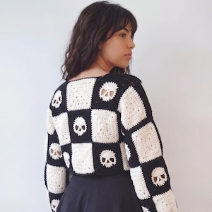 May include: A long-sleeved crochet sweater featuring a checkerboard pattern of alternating black and white squares. Some of the white squares contain a detailed skull motif, while others feature a simple textured square design throughout the garment.