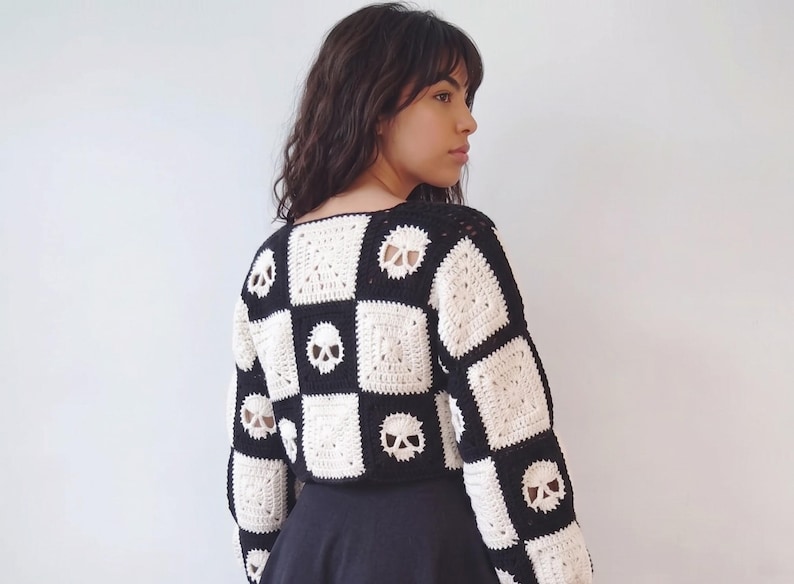 May include: A long-sleeved crochet sweater featuring a checkerboard pattern of alternating black and white squares. Some of the white squares contain a detailed skull motif, while others feature a simple textured square design throughout the garment.