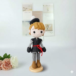 K&ouml;nnte beinhalten: Handgefertigte H&auml;kelpuppe eines Charakters in einem schwarz-grauen Outfit mit rotem G&uuml;rtel. Die Puppe hat hellbraunes Haar, einen schwarzen Hut mit wei&szlig;em Bommel und braune Stiefel. Sie steht auf einem kleinen Holzsockel.