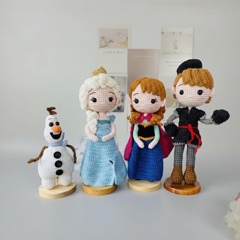 K&ouml;nnte beinhalten: Geh&auml;kelte Puppen von Charakteren aus dem Animationsfilm Frozen. Die Puppen umfassen Olaf den Schneemann, Elsa in einem blauen Kleid, Anna in einem blau-rosa Kleid und Kristoff in einem schwarzen Outfit. Jede Puppe steht auf einem Holzsockel.