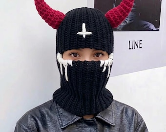 May include: A black crocheted balaclava featuring two large red horns extending from the top. The headgear includes a white embroidered inverted cross on the forehead and white yarn detailing around the eye opening that resembles dripping liquid.