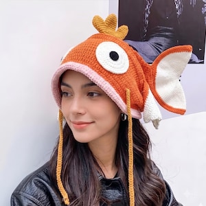 Puede incluir: Un gorro de ganchillo con forma de pez naranja con un borde rosa claro, una aleta dorsal amarilla en la parte superior y grandes ojos blancos con pupilas negras. El gorro presenta una aleta caudal blanca y naranja y dos largas tiras amarillas.