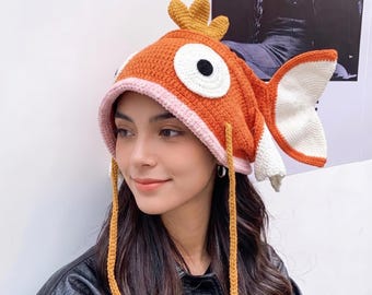 May include: A crochet hat shaped like an orange fish with a light pink brim, large white and black circular eyes, a yellow dorsal fin on top, and a large white and orange tail fin extending from the side, featuring two long yellow chin ties.