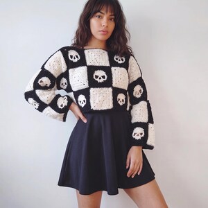 May include: A long-sleeved crochet sweater featuring a black and white checkerboard pattern with skull motifs integrated into the squares, paired with a solid black skater skirt. The sweater has a boxy fit and wide sleeves.