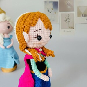 K&ouml;nnte beinhalten: Geh&auml;kelte Puppe einer Figur mit orangefarbenem Haar in einem seitlichen Zopf, die einen rosa Umhang, ein blaues Kleid und einen wei&szlig;en Kragen tr&auml;gt. Die Puppe hat schwarze Knopfaugen und eine Perlenkette.