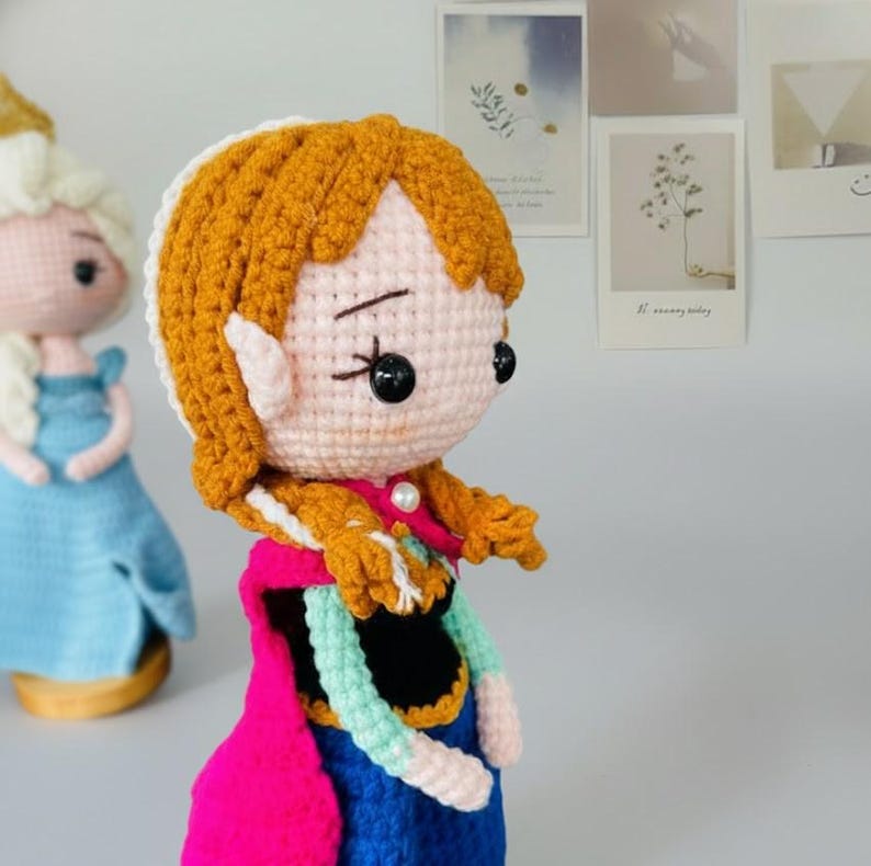 K&ouml;nnte beinhalten: Geh&auml;kelte Puppe einer Figur mit orangefarbenem Haar in einem seitlichen Zopf, die einen rosa Umhang, ein blaues Kleid und einen wei&szlig;en Kragen tr&auml;gt. Die Puppe hat schwarze Knopfaugen und eine Perlenkette.