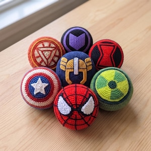 May include: A collection of seven spherical crochet toys featuring various superhero-themed designs. The set includes patterns resembling a red and blue shield with a white star, a red spider web mask, a gold helmet, and other colorful geometric emblems.