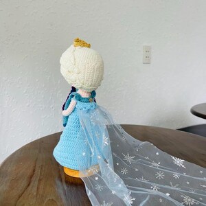K&ouml;nnte beinhalten: Handgefertigte H&auml;kelpuppe mit hellblauem Kleid und wei&szlig;em Kopf mit goldener Krone. Die Puppe hat einen durchsichtigen, hellblauen Umhang mit silbernen Schneeflockenmustern. Die Puppe sitzt auf einer Holzoberfl&auml;che.