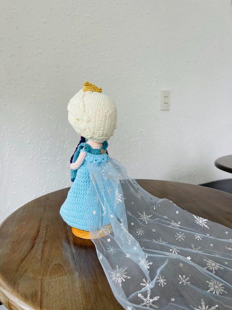 K&ouml;nnte beinhalten: Handgefertigte H&auml;kelpuppe mit hellblauem Kleid und wei&szlig;em Kopf mit goldener Krone. Die Puppe hat einen durchsichtigen, hellblauen Umhang mit silbernen Schneeflockenmustern. Die Puppe sitzt auf einer Holzoberfl&auml;che.