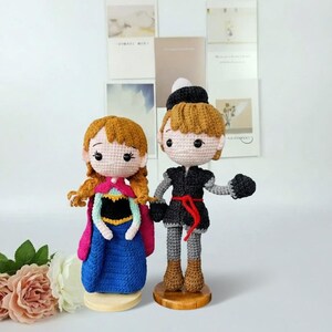 K&ouml;nnte beinhalten: Zwei handgefertigte H&auml;kelpuppen, eine in einem blauen Kleid mit pinkfarbenem Umhang und die andere in einem schwarzen Outfit mit rotem G&uuml;rtel. Beide Puppen haben hellbraunes Haar und stehen auf Holzsockeln. Die Puppen stehen vor einem wei&szlig;en Hintergrund.