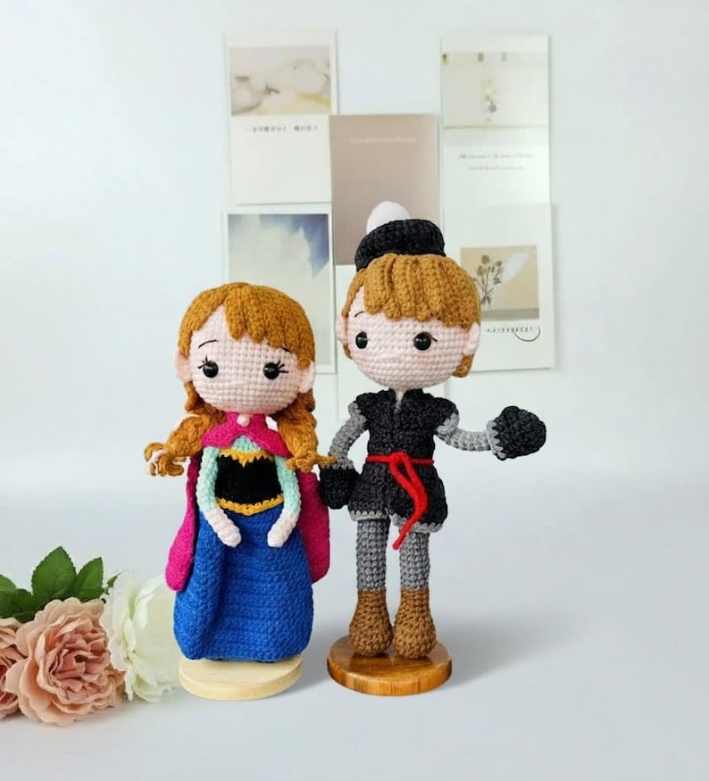 K&ouml;nnte beinhalten: Zwei handgefertigte H&auml;kelpuppen, eine in einem blauen Kleid mit pinkfarbenem Umhang und die andere in einem schwarzen Outfit mit rotem G&uuml;rtel. Beide Puppen haben hellbraunes Haar und stehen auf Holzsockeln. Die Puppen stehen vor einem wei&szlig;en Hintergrund.