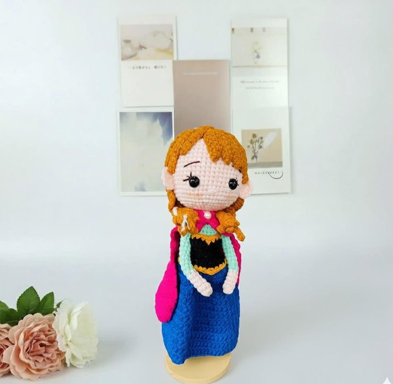 K&ouml;nnte beinhalten: Handgemachte H&auml;kelpuppe einer Figur mit orangefarbenem Haar, die ein blaues Kleid, einen rosa Umhang und ein schwarzes Oberteil tr&auml;gt. Die Puppe hat einen hellen Hautton und wird auf einem Holzst&auml;nder ausgestellt. Der Hintergrund umfasst dekorative Karten.