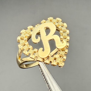 May include: A gold ring with the letter "R" in a heart-shaped design. The letter is in a cursive font, and the heart has a textured border. The ring is held by tweezers against a dark background. The ring is likely made of gold.