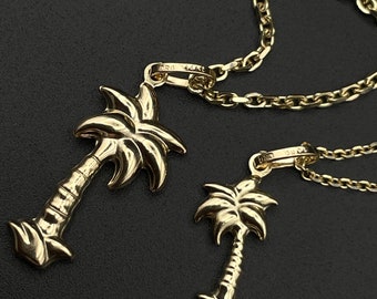 May include: A gold chain necklace with two palm tree pendants. The larger pendant is on the left and the smaller pendant is on the right. The chain is a delicate, cable chain.