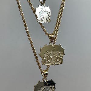 Pu&ograve; includere: Tre collane color oro con ciondoli di Betty Boop. I ciondoli presentano una superficie strutturata e l'immagine del personaggio dei cartoni animati. Le catene sono in stile corda. Le collane sono mostrate su uno sfondo neutro.