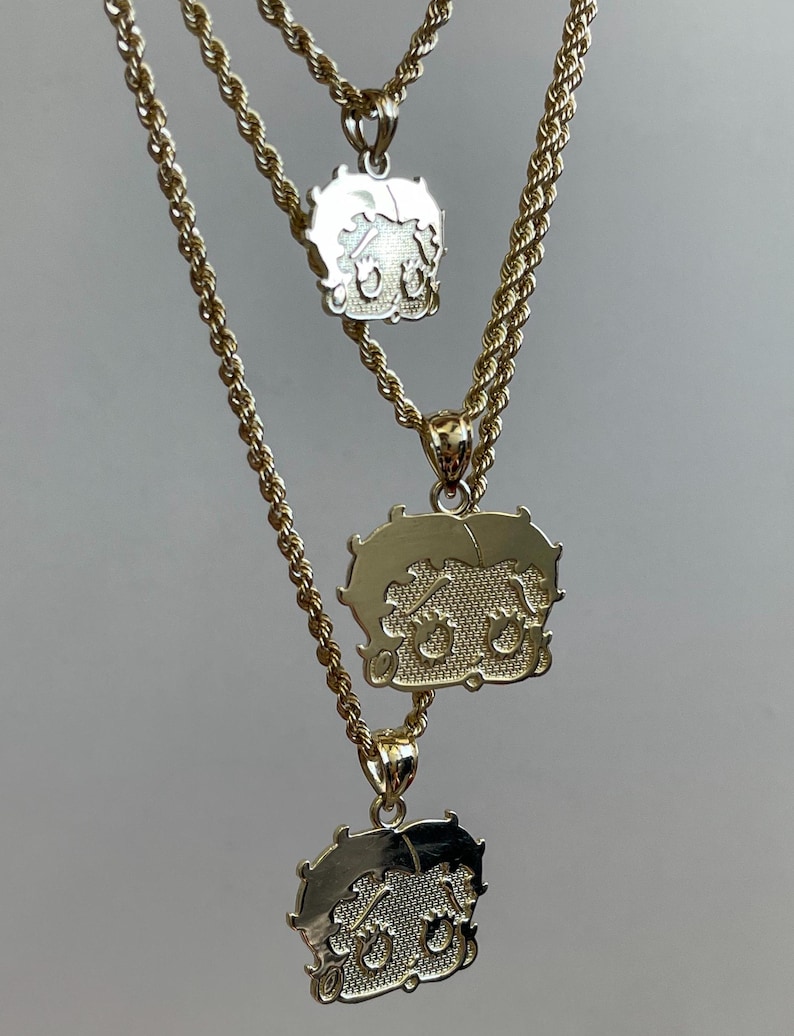 Pu&ograve; includere: Tre collane color oro con ciondoli di Betty Boop. I ciondoli presentano una superficie strutturata e l'immagine del personaggio dei cartoni animati. Le catene sono in stile corda. Le collane sono mostrate su uno sfondo neutro.