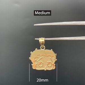 Pu&ograve; includere: Un ciondolo color oro a forma di personaggio dei cartoni animati, tenuto da una pinzetta su sfondo scuro. Il ciondolo misura circa 20 mm di larghezza. La parola "Medium" &egrave; visualizzata sopra il ciondolo.