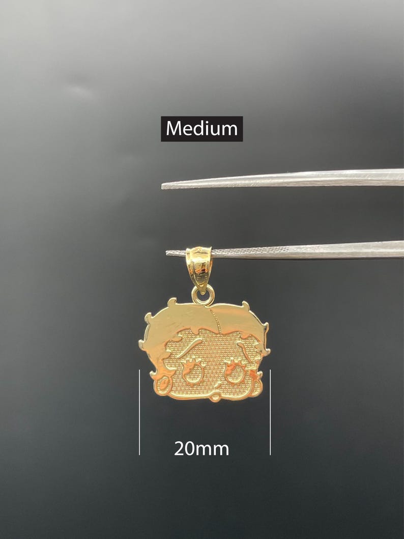 Pu&ograve; includere: Un ciondolo color oro a forma di personaggio dei cartoni animati, tenuto da una pinzetta su sfondo scuro. Il ciondolo misura circa 20 mm di larghezza. La parola "Medium" &egrave; visualizzata sopra il ciondolo.
