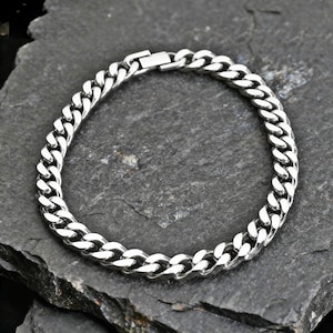 Puede incluir: Un brazalete de cadena de color plateado. El brazalete está hecho de eslabones entrelazados y tiene un cierre seguro. La joya se exhibe sobre una superficie oscura y texturizada.