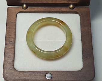 May include: A smooth, circular ring crafted from translucent pale green jade with subtle amber-colored inclusions. The ring is displayed resting flat inside a square, dark brown wooden box featuring a white, soft-textured interior lining.