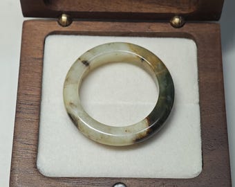 May include: A smooth, circular band ring crafted from polished stone, featuring a variegated color palette of translucent off-white, deep forest green, and dark brown inclusions. The ring is presented inside a square, dark-toned wooden box.
