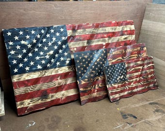 Può includere: Tre bandiere americane in legno di diverse dimensioni, ognuna caratterizzata da una superficie ondulata e increspata. Le bandiere sono tinte in rosso, bianco e blu, con una finitura in legno bruciato che mette in risalto le venature.