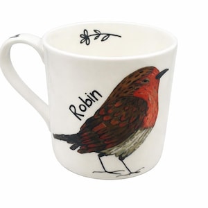 May include: White ceramic mug featuring a detailed illustration of a robin bird with red breast and brown wings. The word "Robin" is written in black script. A small sprig of leaves is near the rim. The mug has a curved handle.