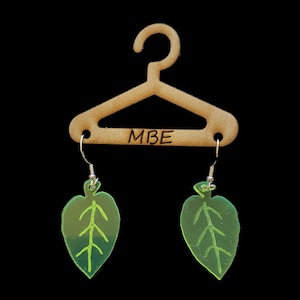 以下が含まれることがあります： 木製のハンガーに「MBE」と刻印された、銀色のフックに吊るされた緑の葉の形をしたイヤリングのペアです。