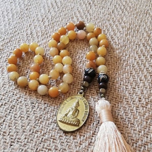May include: A yellow and brown beaded mala necklace with a gold pendant featuring a seated Buddha. The necklace has a tassel at the end.