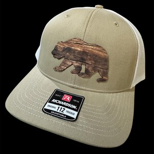 May include: A tan trucker hat with a brown bear applique. The hat has a curved peak, white mesh back, and an adjustable strap. The Richardson brand label is visible on the front.