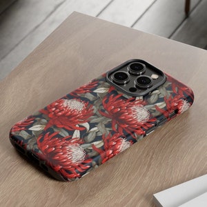 Puede incluir: Una funda de teléfono negra con un diseño floral con flores rojas y hojas verdes. La funda tiene una protuberancia de cámara elevada con tres lentes de cámara negras.
