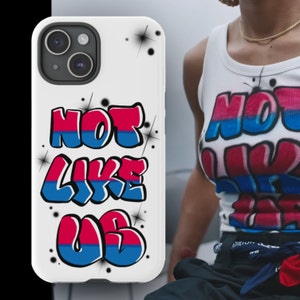 May include: White iPhone case with the words "NOT LIKE US" in red and blue graffiti style. The case has a white background with black starburst accents. The image also shows a person wearing a white tank top with the same text.
