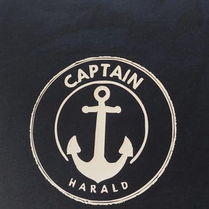 Könnte beinhalten: Marineblaues T-Shirt mit weißem, kreisförmigem Aufdruck. Der Aufdruck zeigt einen Anker in einem Kreis mit dem Wort "CAPTAIN" darüber und "HARALD" darunter. Das Design hat einen leicht verblichenen, Vintage-Look.
