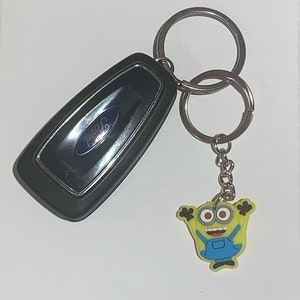 May include: A black Ford car key with a silver key ring and a yellow Minion keychain. The Minion is wearing blue overalls and has red eyes. The key has the Ford logo.