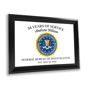 Può includere: Una targa incorniciata in nero con sfondo bianco. La targa presenta il sigillo del Federal Bureau of Investigation (FBI) e il testo "34 YEARS OF SERVICE Andrew Wilson FEDERAL BUREAU OF INVESTIGATION EST. MAY 23, 1990".