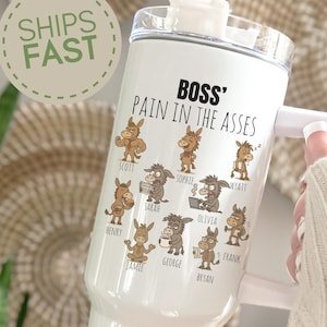 May include: White tumbler with a handle and straw, featuring the text "BOSS' PAIN IN THE ASSES" and cartoon donkeys with names. The tumbler has a clear lid and a white handle. A green and white "SHIPS FAST" sticker is visible.