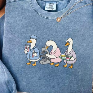 May include: Blue jean colored sweatshirt featuring a graphic of three cartoon ducks in lab coats, one holding test tubes, one using a microscope, and one holding a tray of petri dishes. The text "BLUE JEAN" is at the bottom.