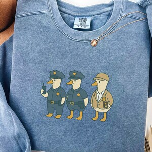 Może przedstawiać: Niebieska bluza dżinsowa z grafiką przedstawiającą trzy kaczki z kresk&oacute;wek ubrane w mundury policjant&oacute;w. Kaczki noszą czapki i mundury policyjne. Bluza ma okrągły dekolt i długie rękawy. Tekst "BLUE JEAN" znajduje się na dole.